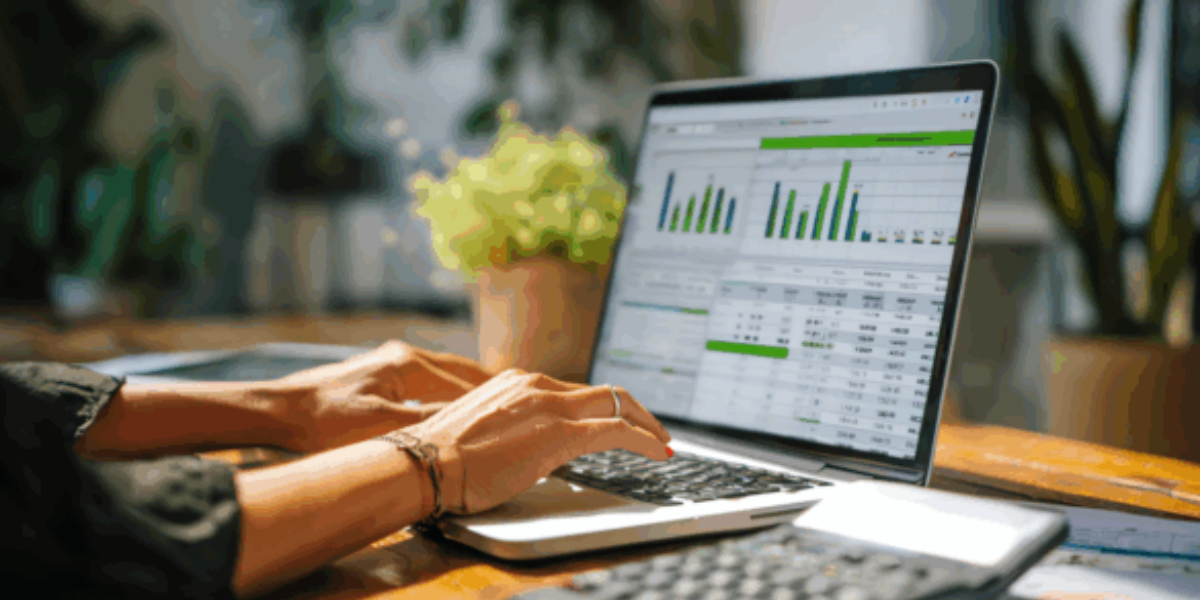 Close up of hands typing on a laptop displaying green financial charts and spreadsheets, with a calculator and documents on the desk.