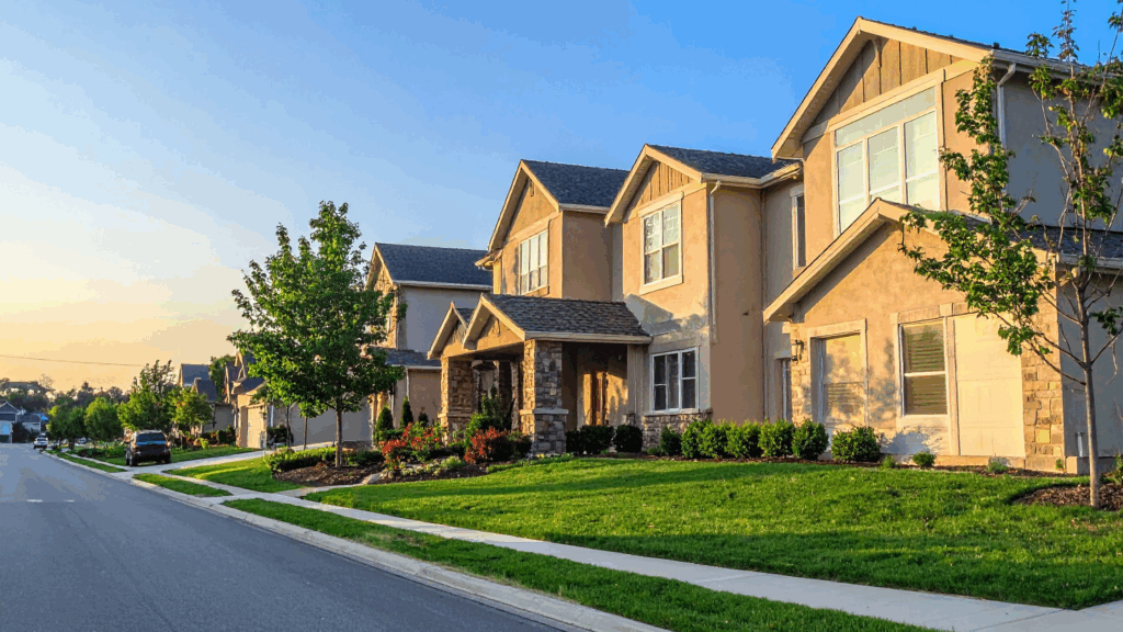 Suburban street lined with modern two story homes, green lawns, and trees at sunset, showing a peaceful residential neighborhood.
