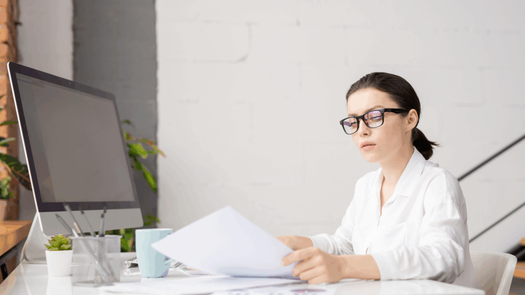 A woman wearing glasses sits at a desk in a modern office, reviewing documents with a computer monitor, coffee mug, and office supplies beside her.
