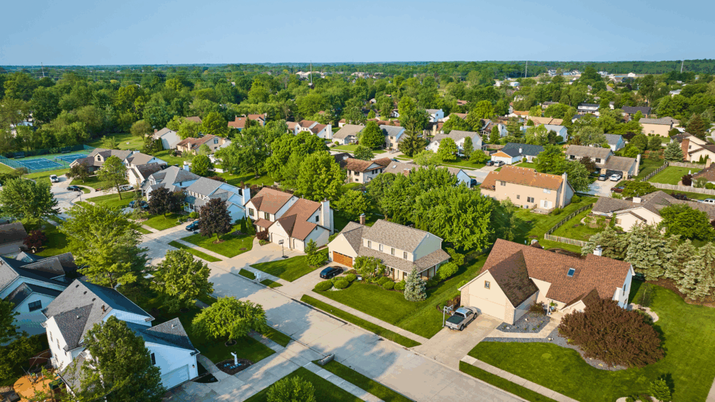 Aerial view of a suburban neighborhood with single family homes, tree lined streets, and well maintained yards on a sunny day.