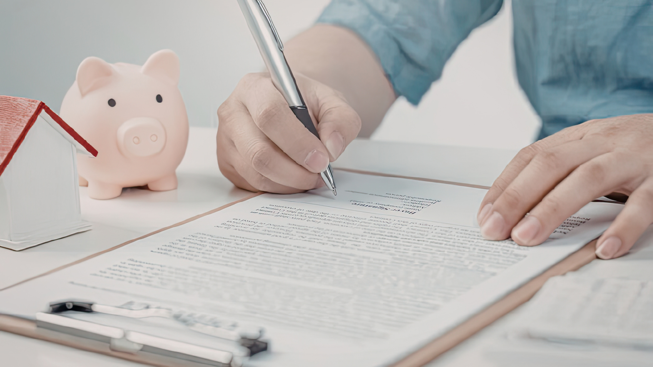 Person signing real estate documents with a piggy bank and small house model on the table
